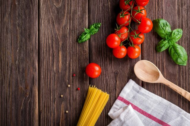 Des tomates, des spaghettis, du basilic, ainsi que du poivre blanc et rouge, une cuillère en bois et un chiffon sont présentées sur une table en bois.